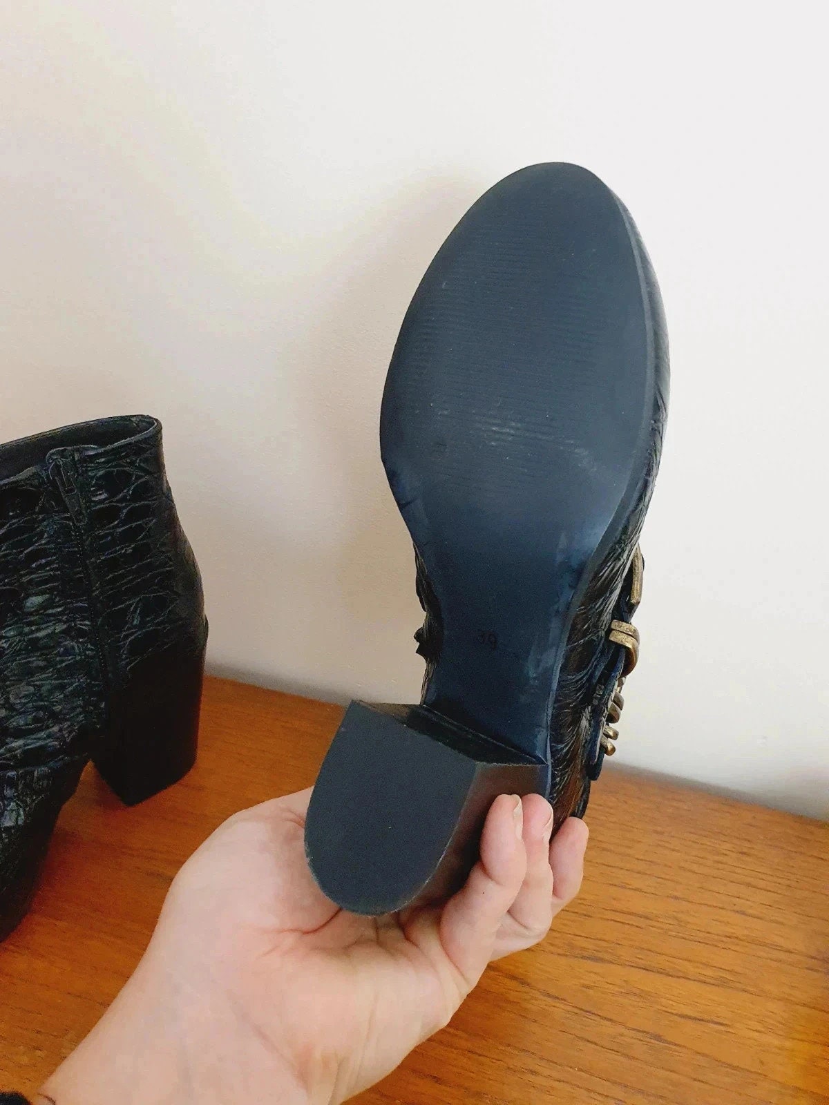 Person holding a black boot with a white wall and wooden floor in the background