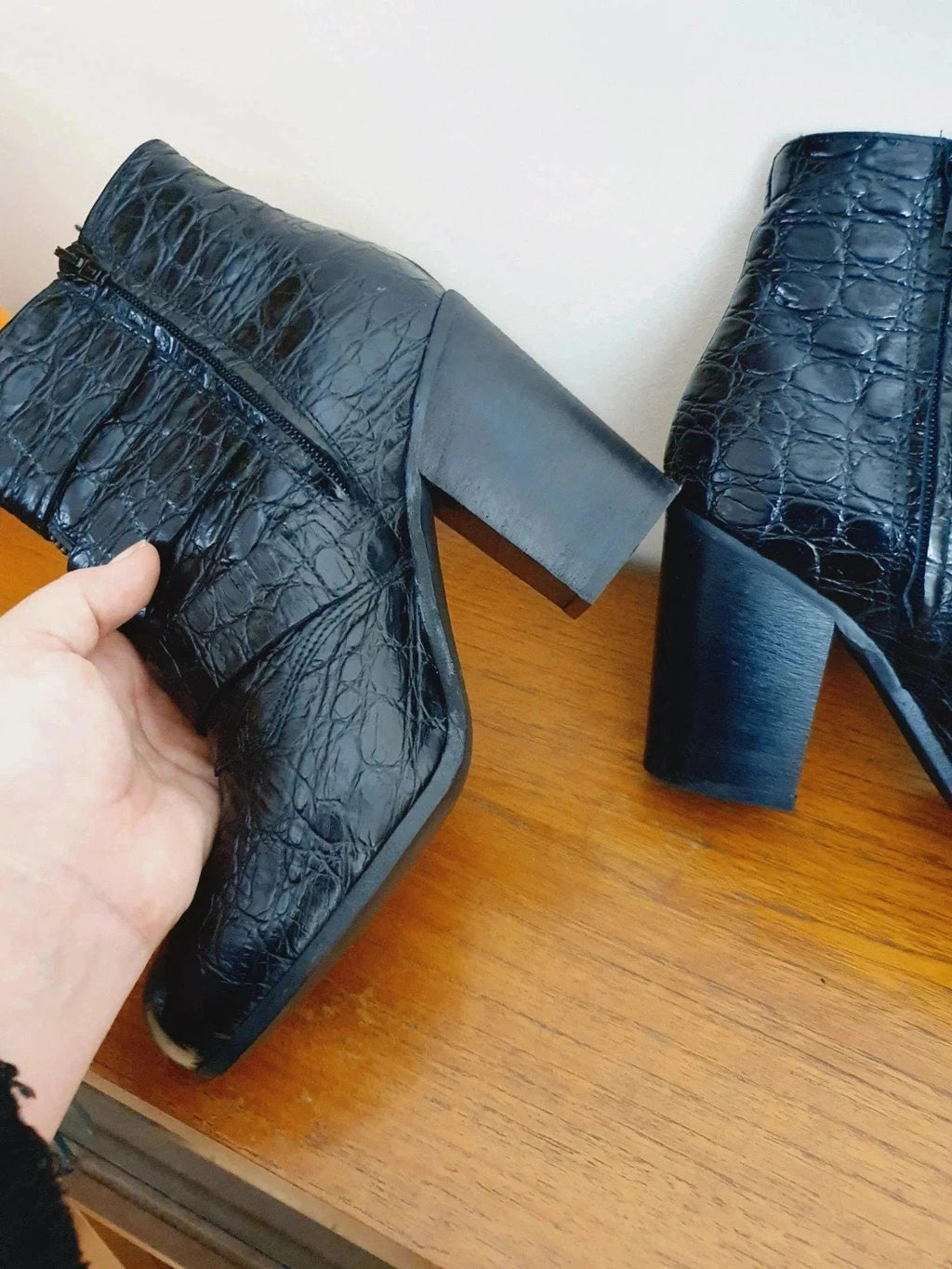 Black crocodile-patterned high-heeled boots on a wooden surface with a hand holding one boot.
