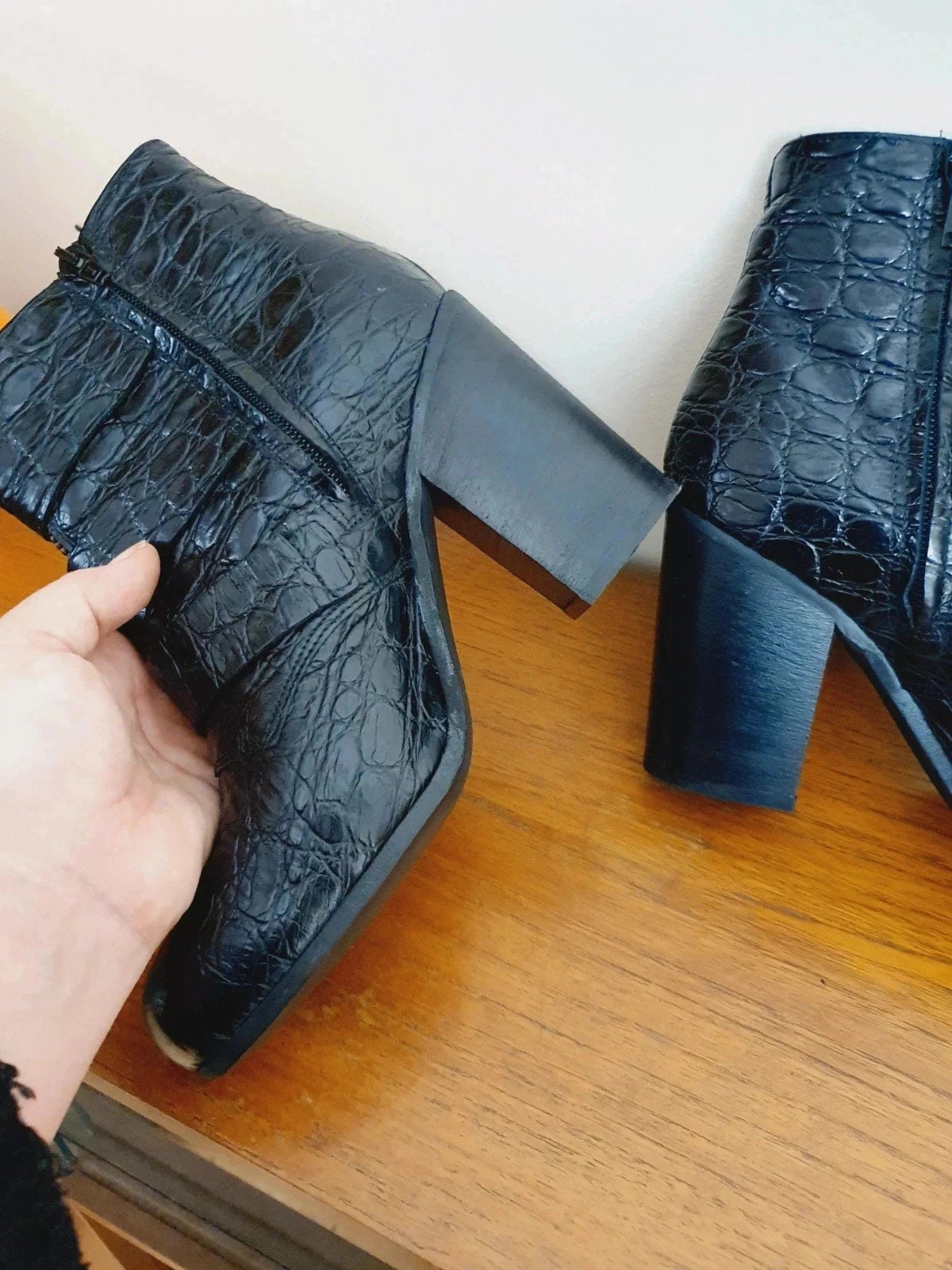 Black crocodile-patterned high-heeled boots on a wooden surface with a hand holding one boot.