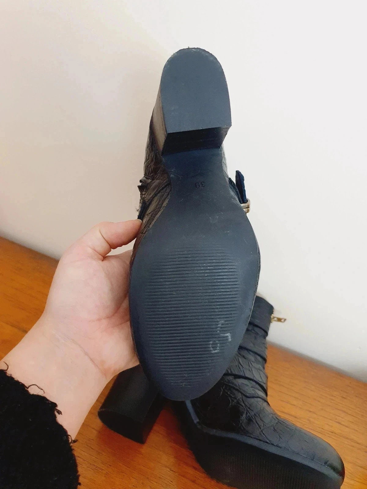 Black boot with a textured sole held by a hand on a wooden surface.