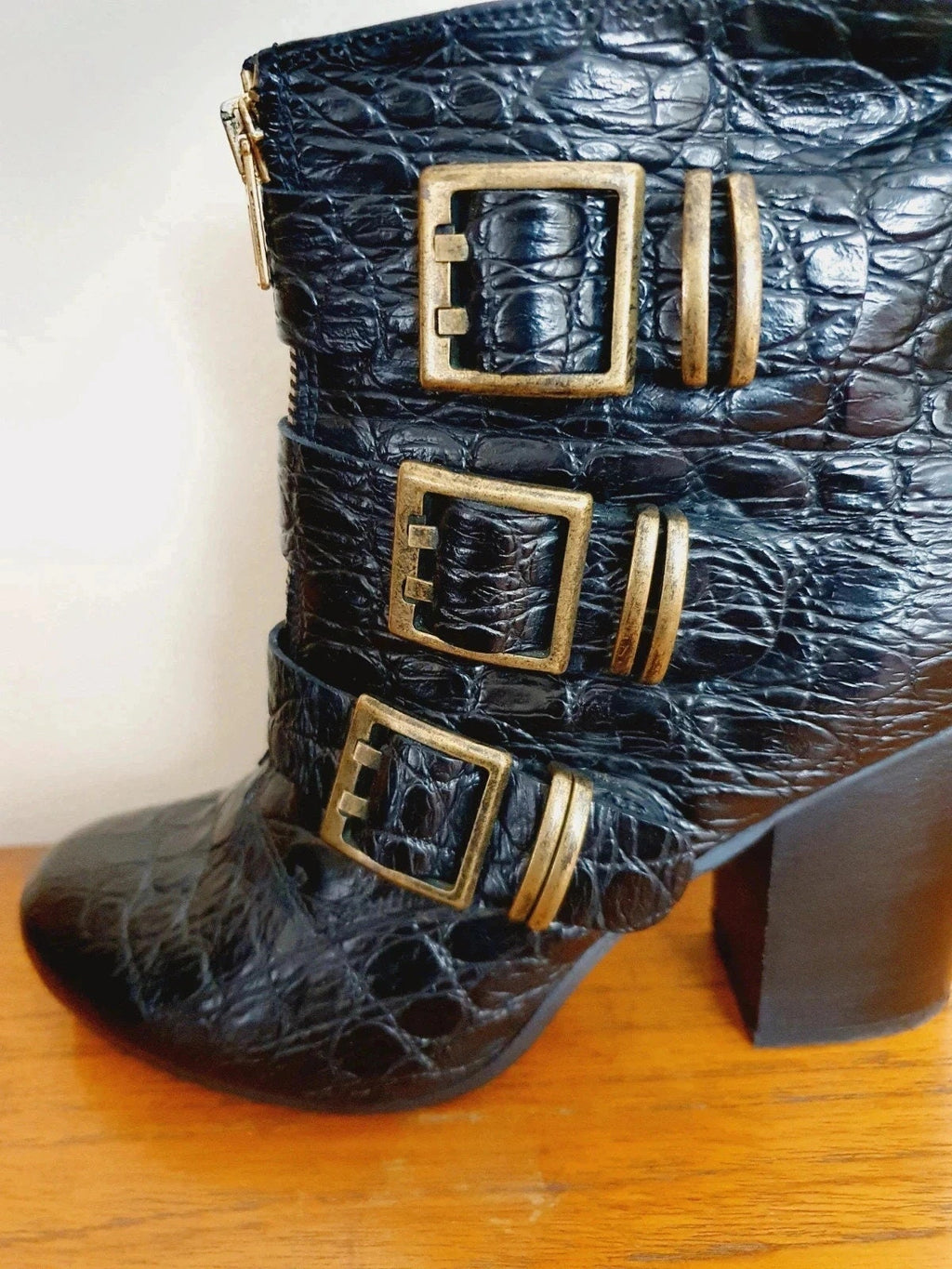 Black crocodile-patterned boot with gold buckles on a wooden surface.