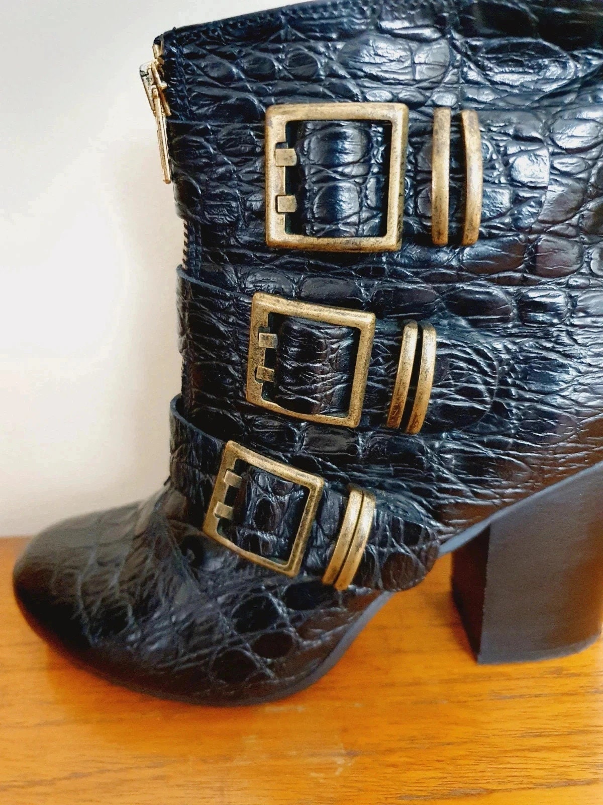 Black crocodile-patterned boot with gold buckles on a wooden surface.